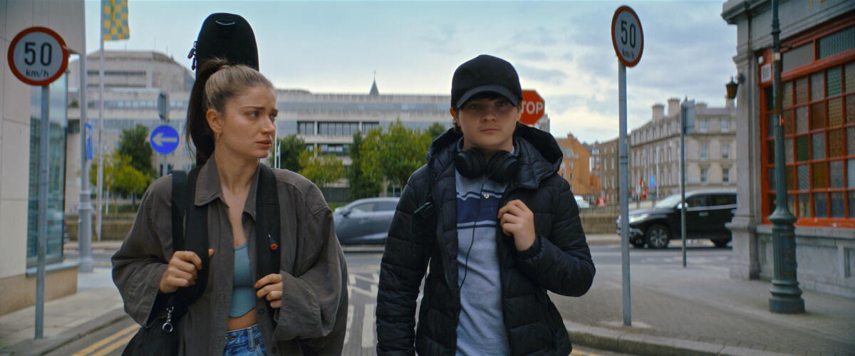 A woman carrying a guitar walking on a city street next to her son, who wears headphones around his neck.