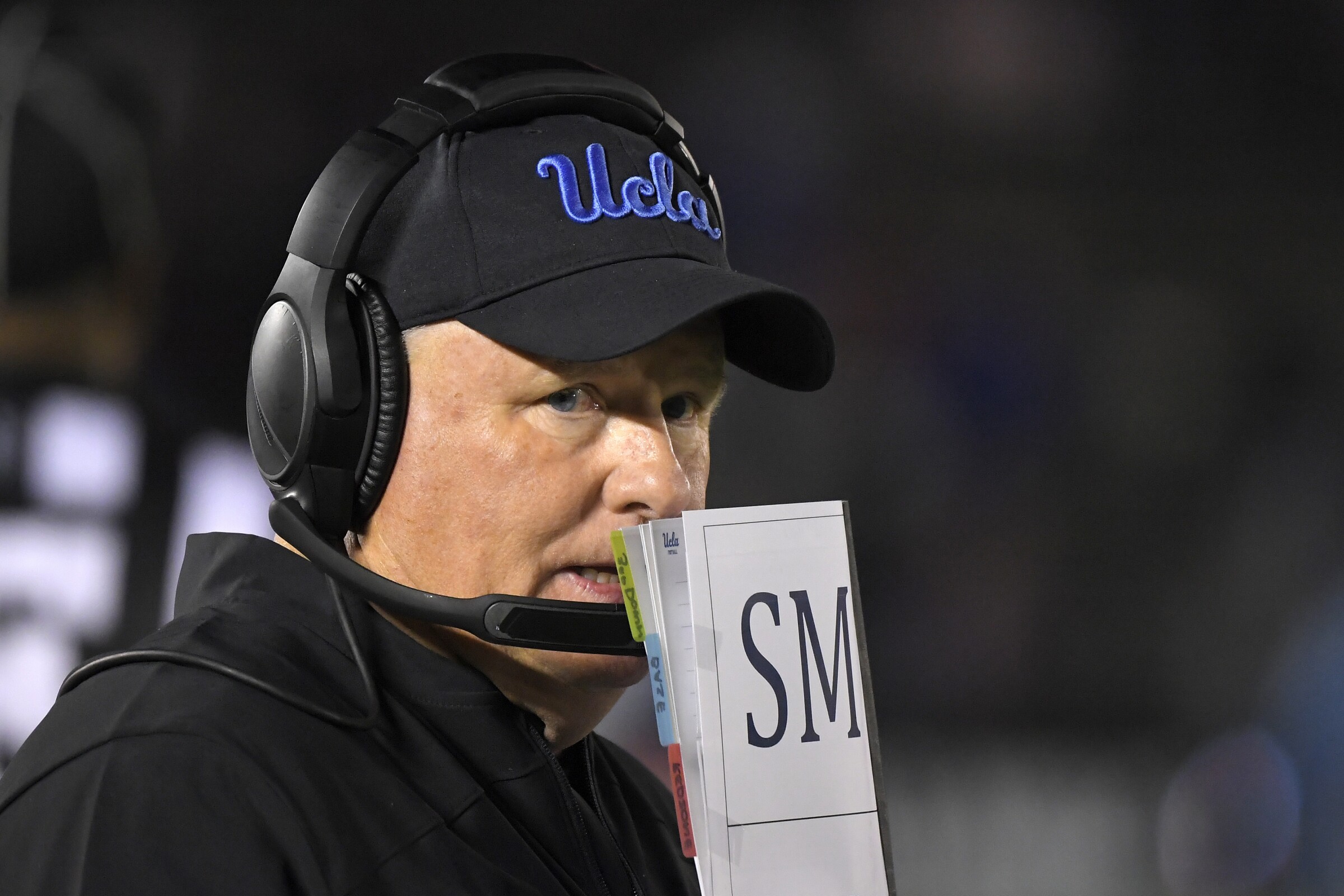 UCLA head coach Chip Kelly covers his mouth with a playbook while standing on the sideline