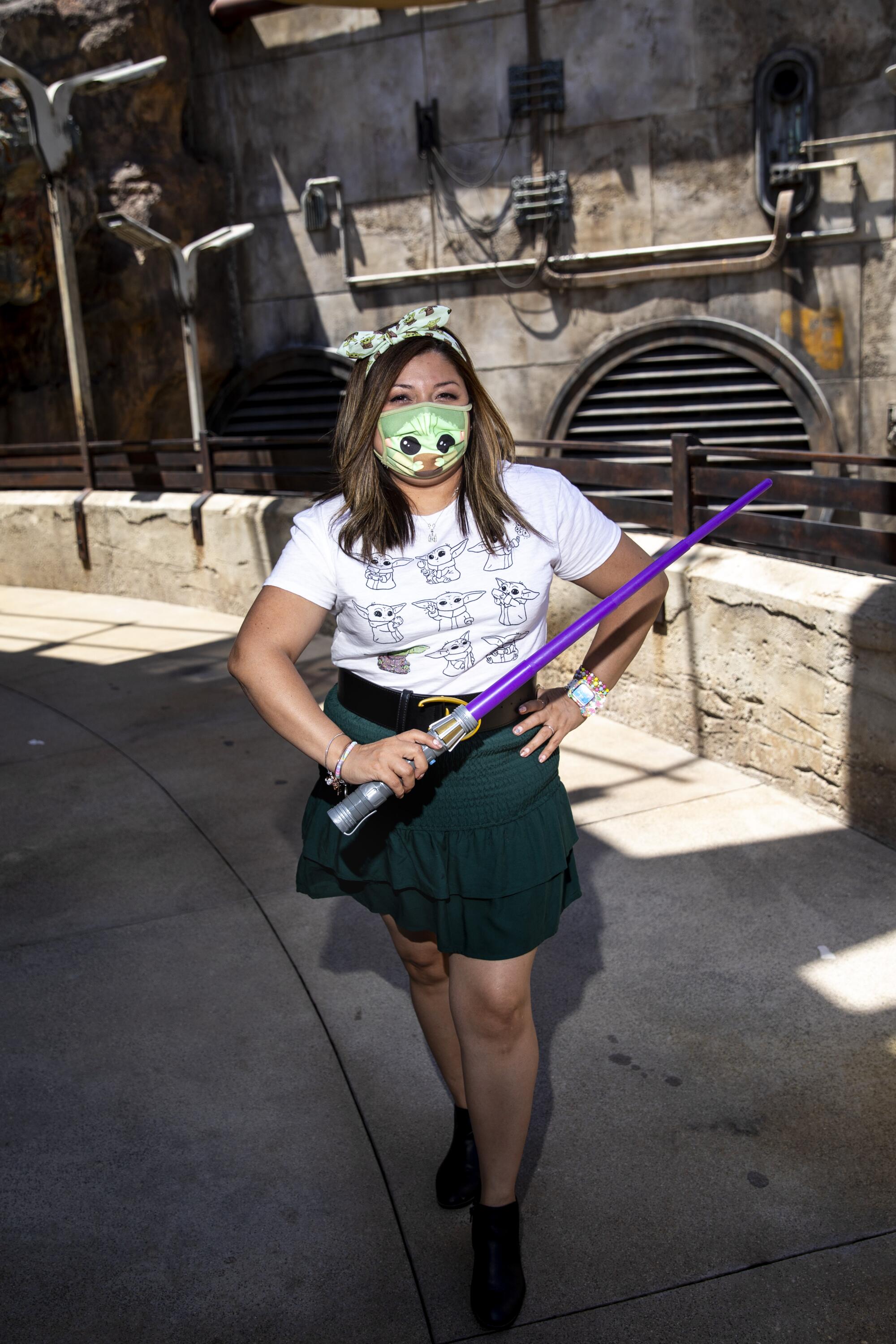 A woman in a baby Yoda mask and shirt holds a purple lightsaber.