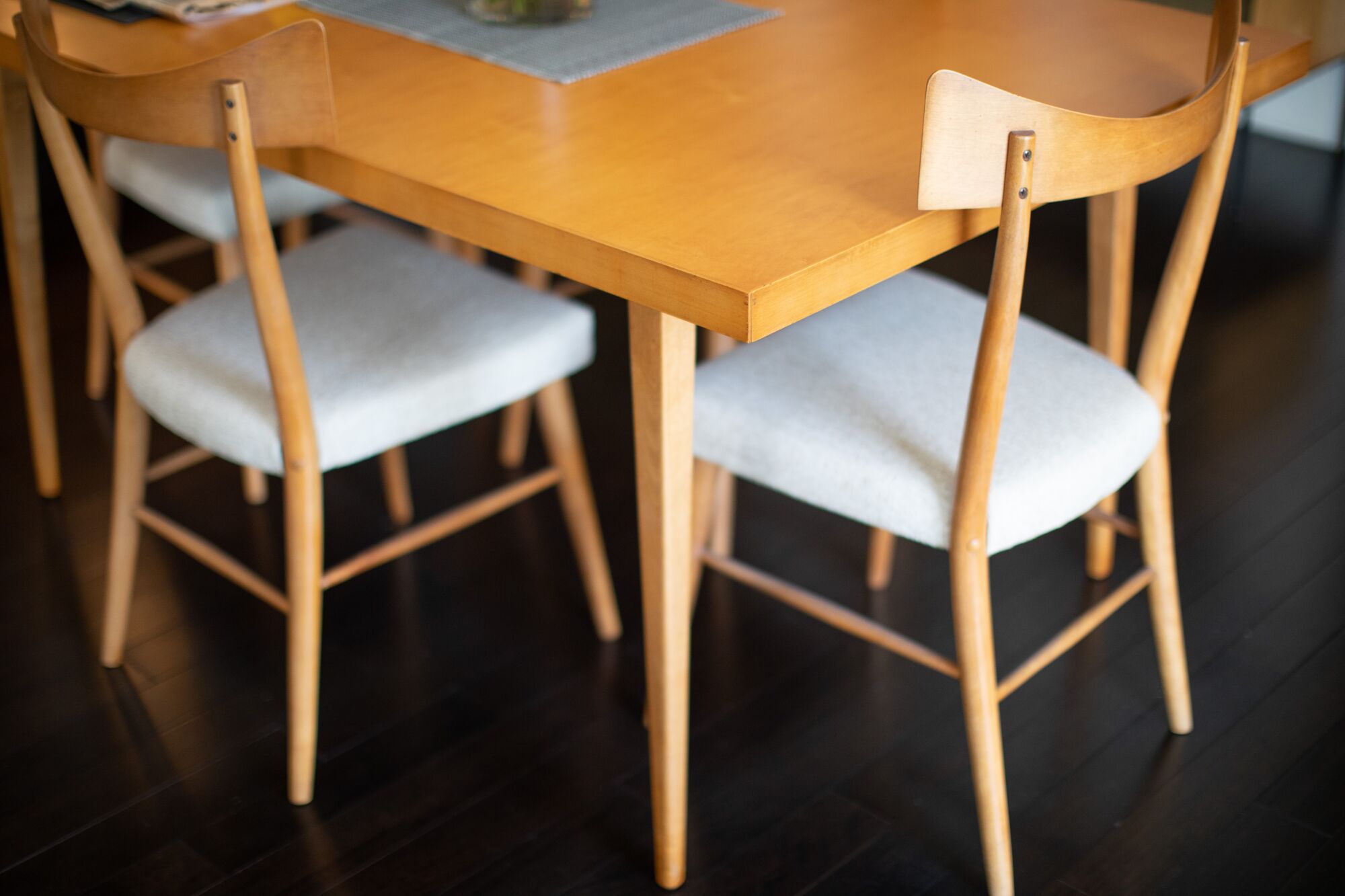 A photo taken at an angle reveals a corner of a wood dining table and curved back dining chairs in the Modernist style