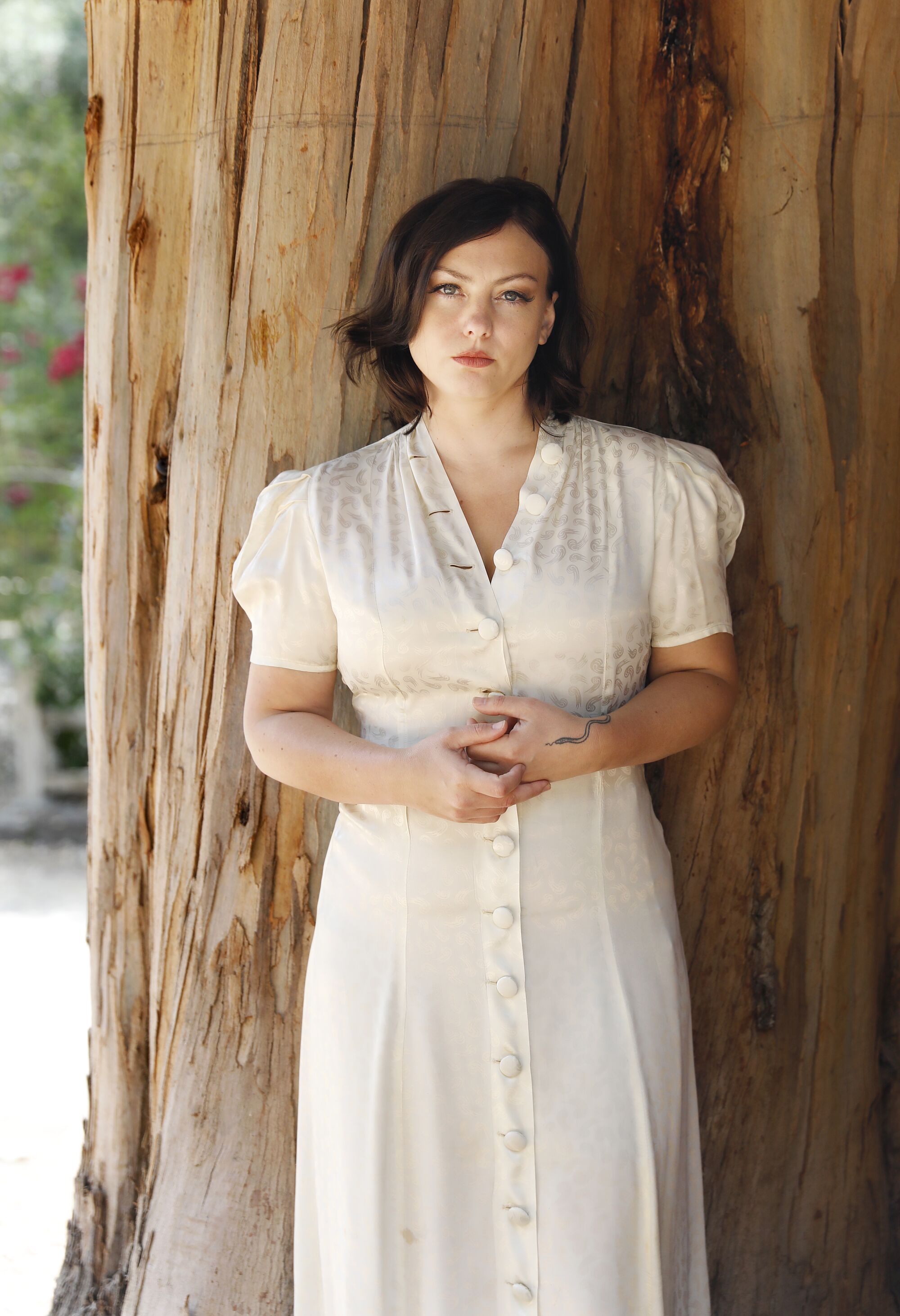 A woman in a white dress leans on a tree trunk