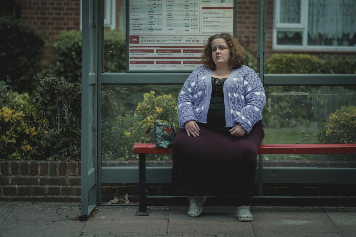 Una mujer con un vestido y un suéter morado sentada en el banco de una parada de autobús.
