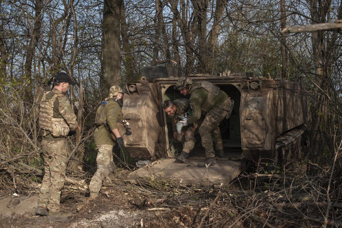 Military medics help a wounded soldier out of a vehicle in a forested area.