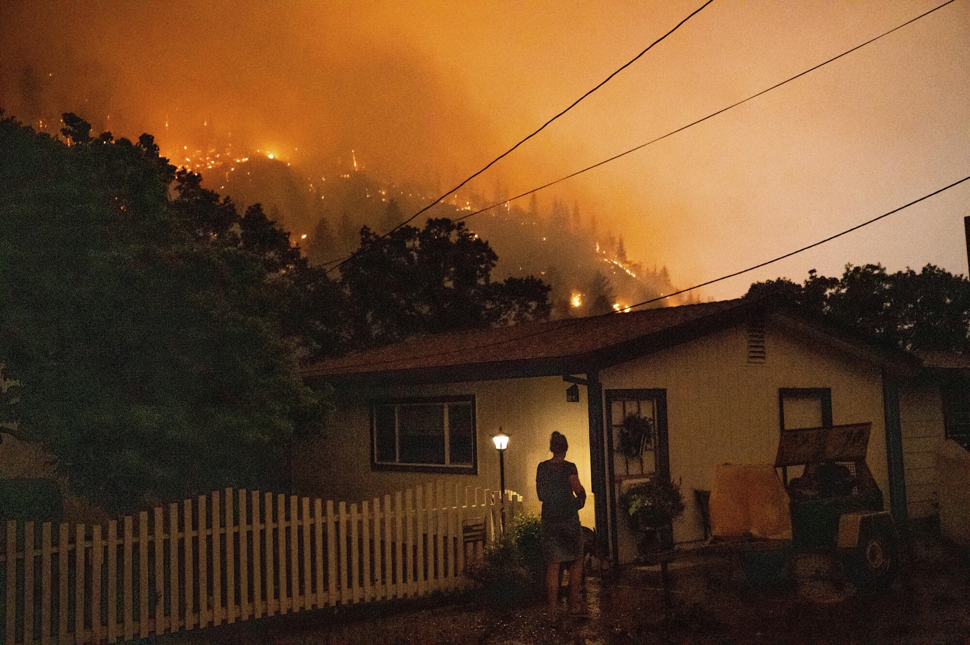 A woman outside a house framed by a burning ridge nearby.