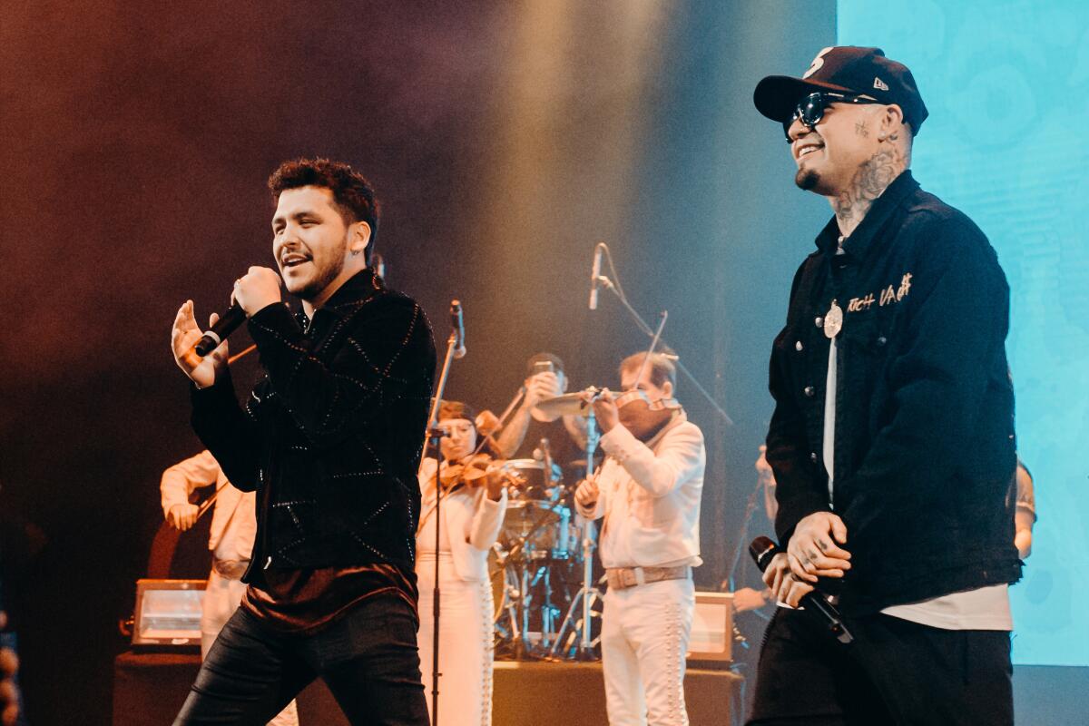 On stage, with violin players in the background, Christian Nodal and Gera MX hold microphones.