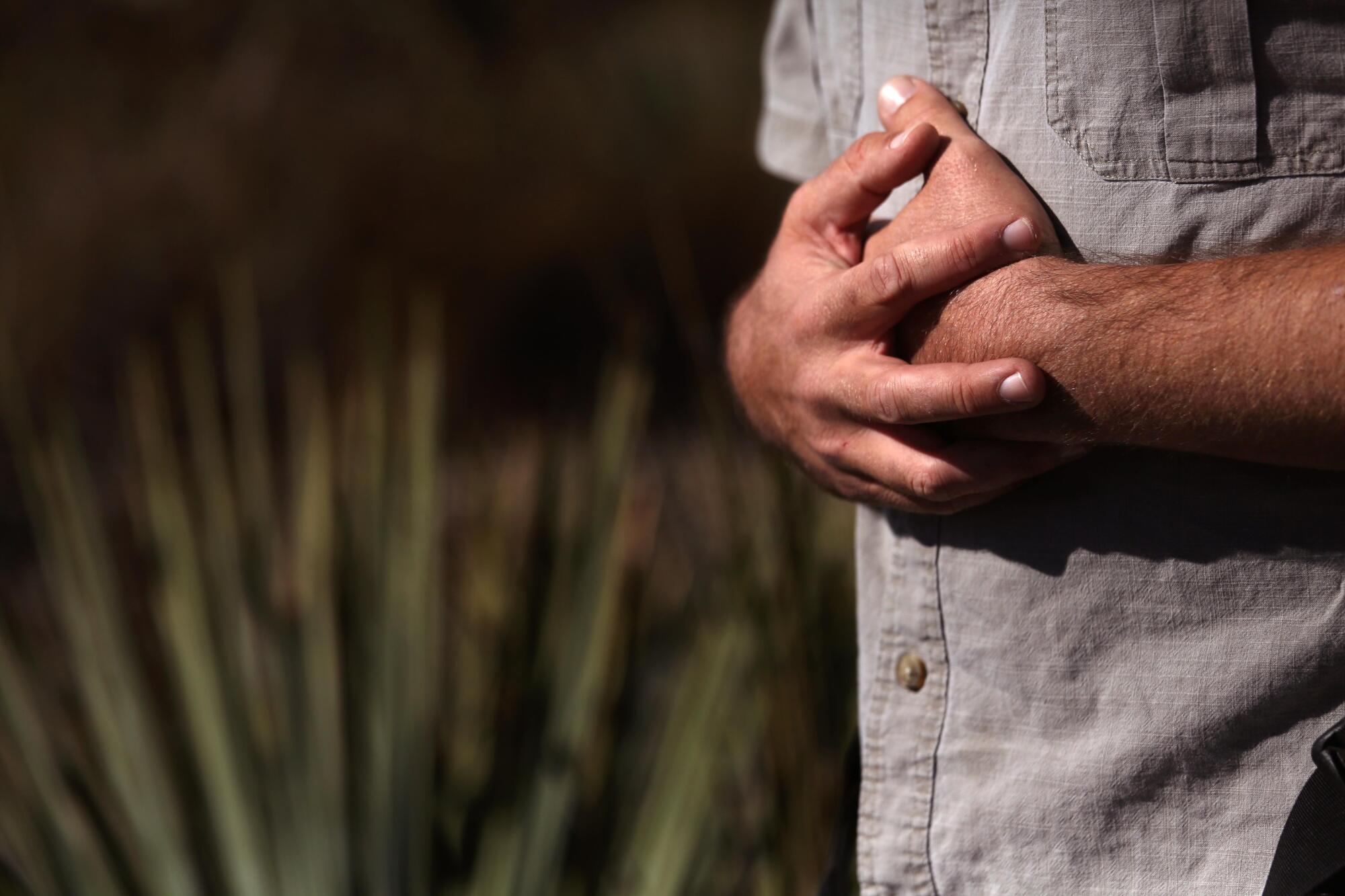 A man, visible from chest to waist, stands in front of a spiky plant, hands folded over his midsection