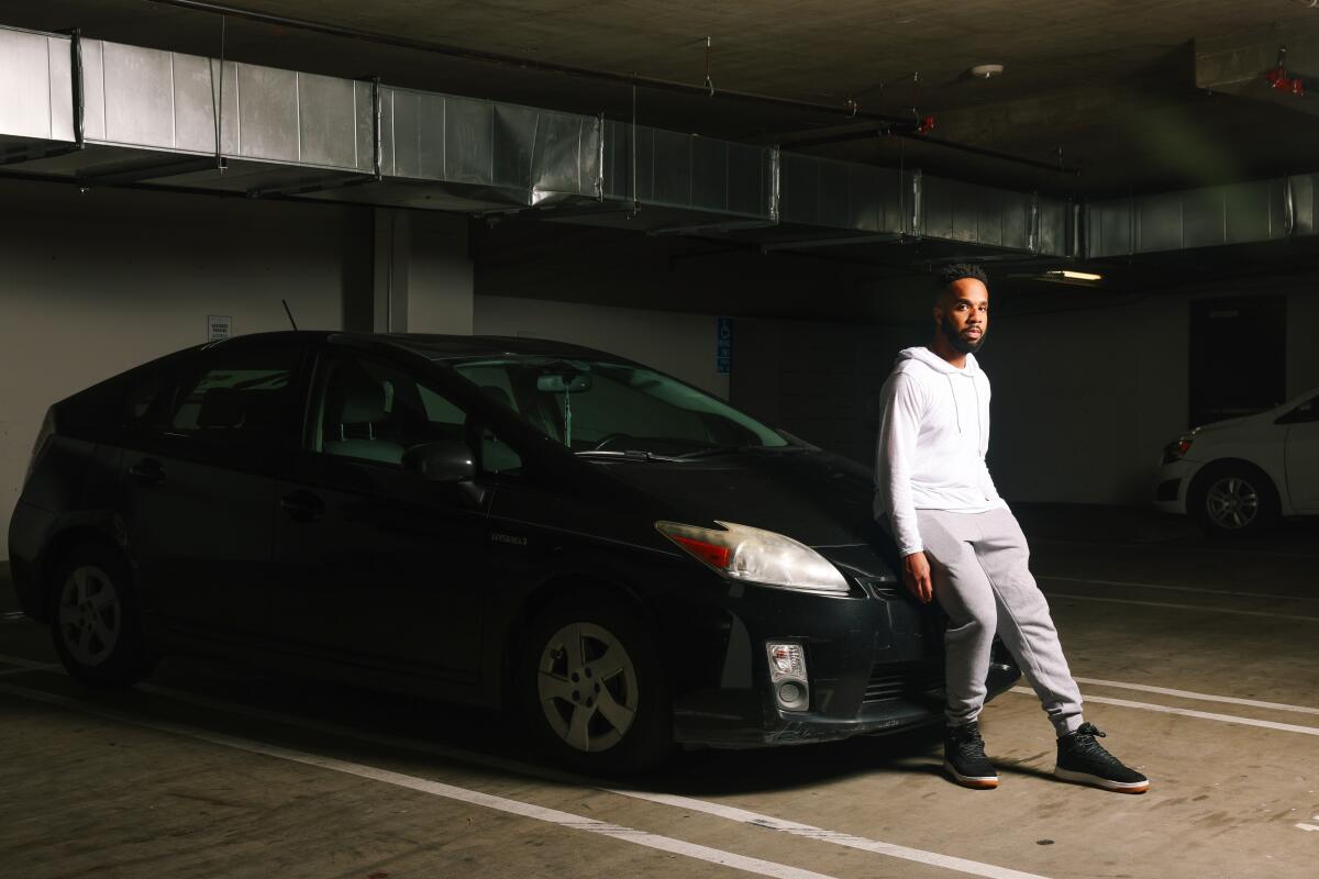 A man wearing a white shirt and gray pants leans against the hood of a black Toyota Prius inside a parking garage.