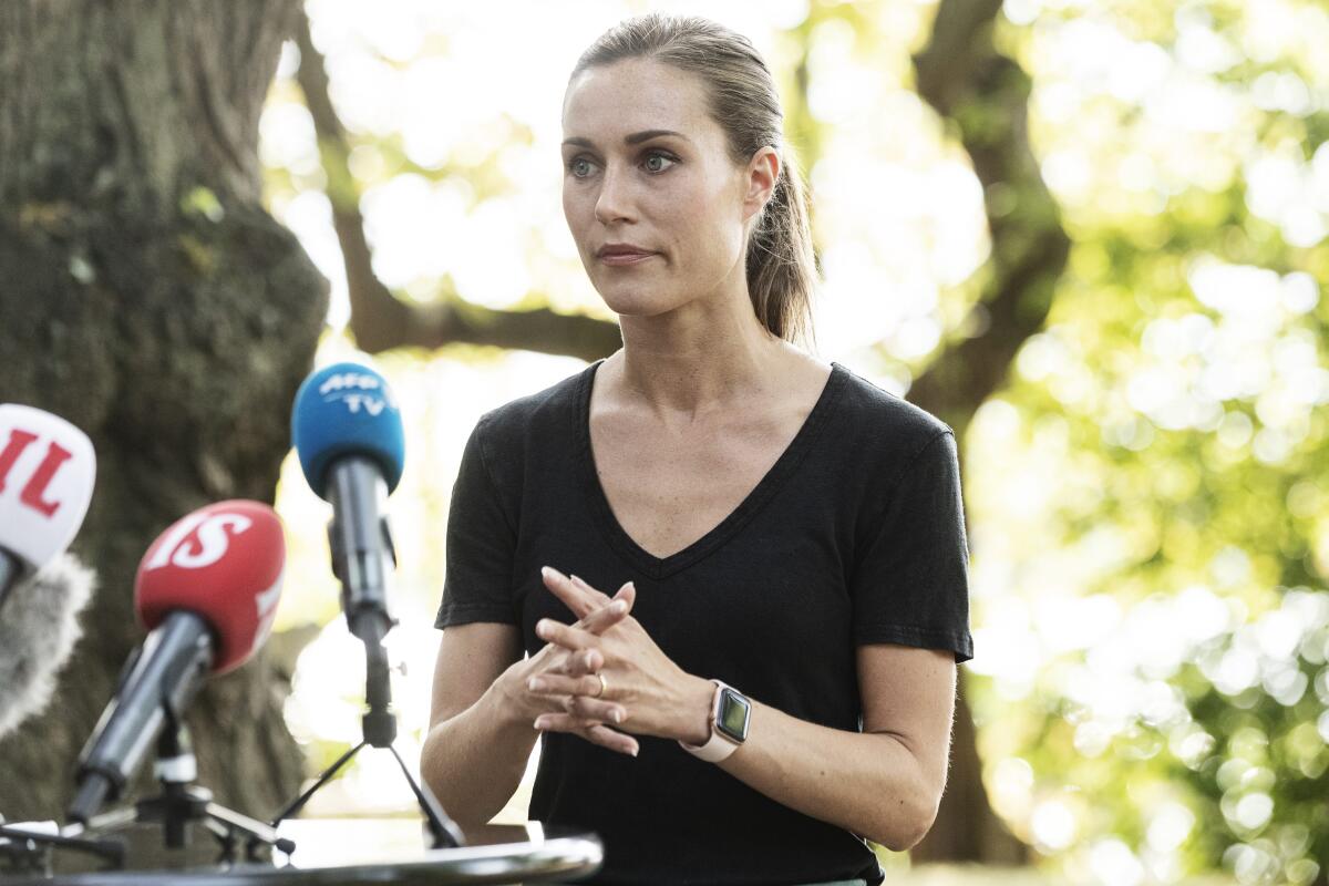 Prime Minister of Finland Sanna Marin stands with her fingers laced in front of microphones outdoors.