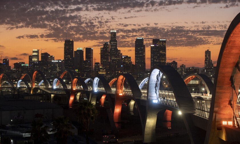 Clouds float over city skyscrapers as sunset turns the sky peach. In the foreground the arches of a bridge are lighted up.