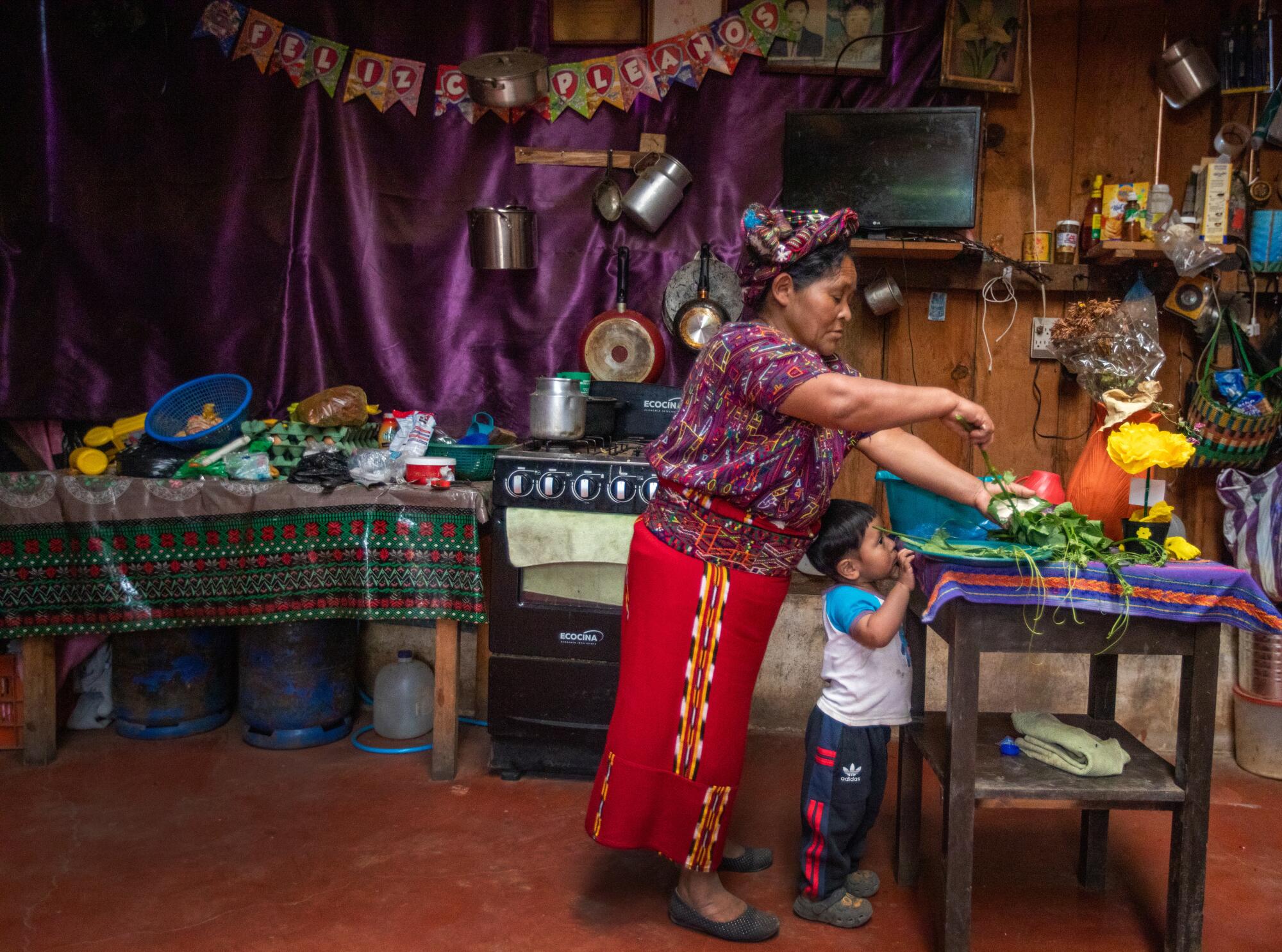 A woman in colorful, traditional dress prepares a traditional Mayan Ixil dish. A boy stands in front of her, watching.
