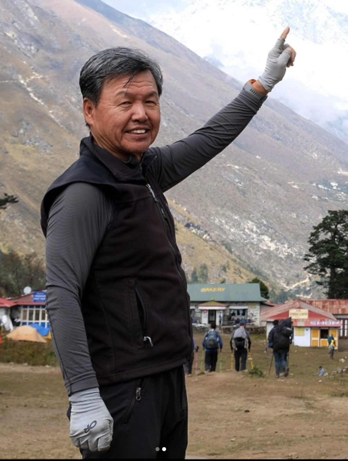 A man standing in an open area in front of a mountain points at the sky.