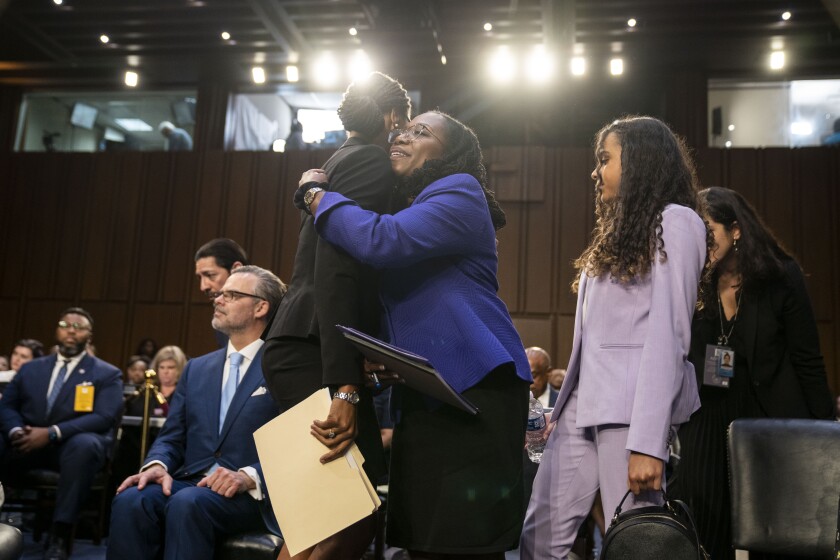 A Black woman wears a blue jacket and hugs another woman in a wood-paneled room