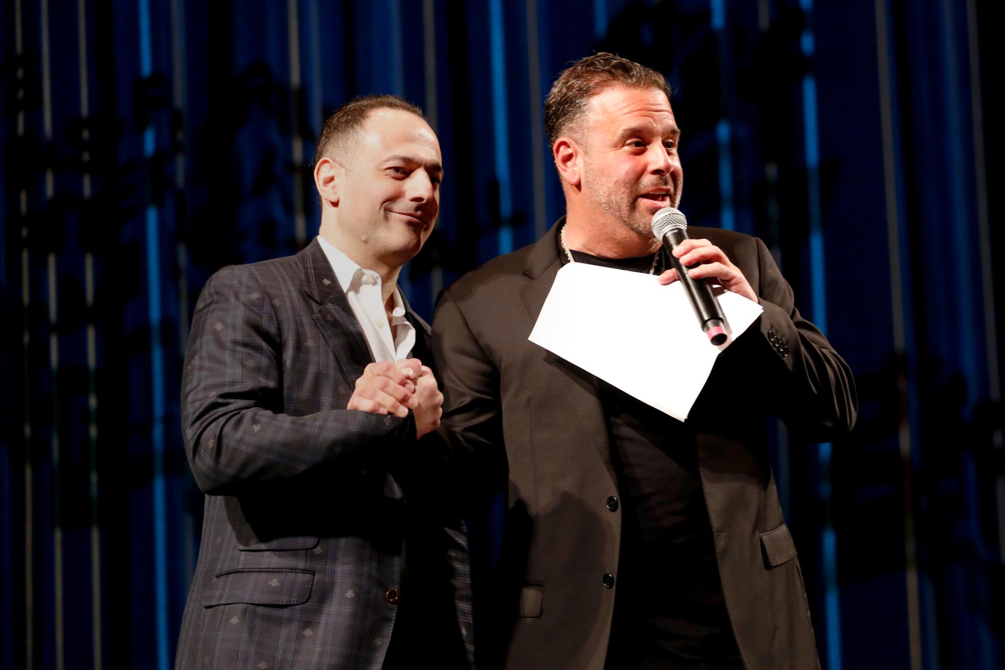 Joel Cohen and Randall Emmett clasp hands onstage as Emmett speaks into a microphone