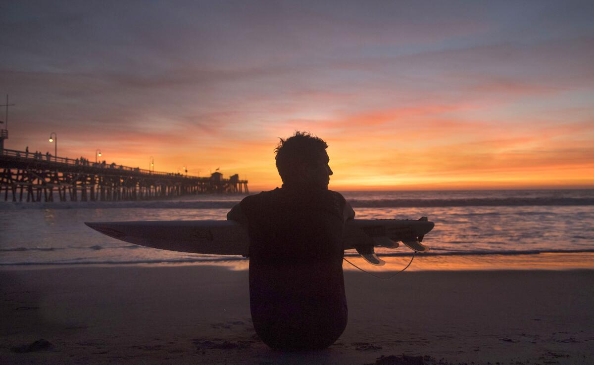 A surfer sits on the beach, holding his board, and watches the sunset.