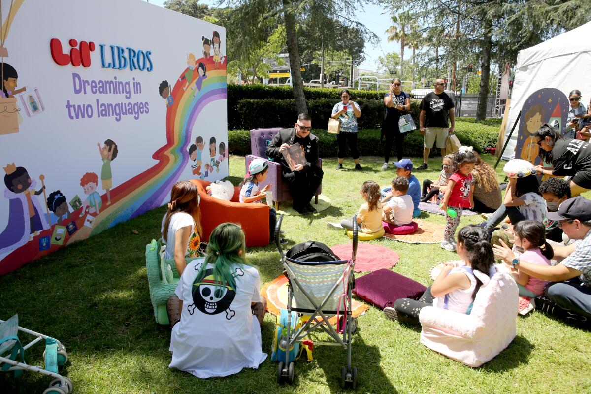 Children gather on a grassy area to listen to a story being read. A sign saying "Lil' Libros" is in the background