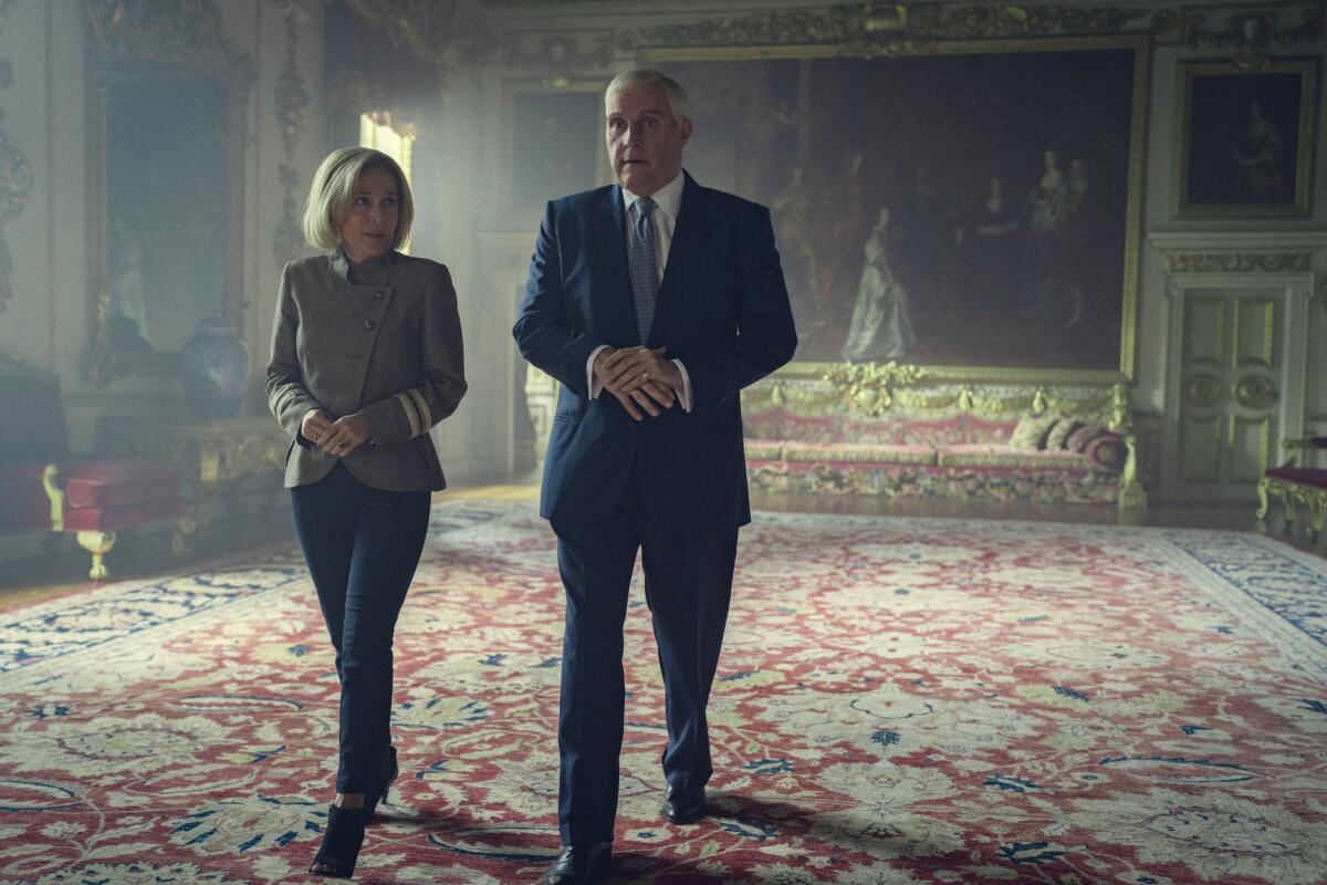 A man and a woman walk on a patterned carpet in an opulent room in "Scoop."