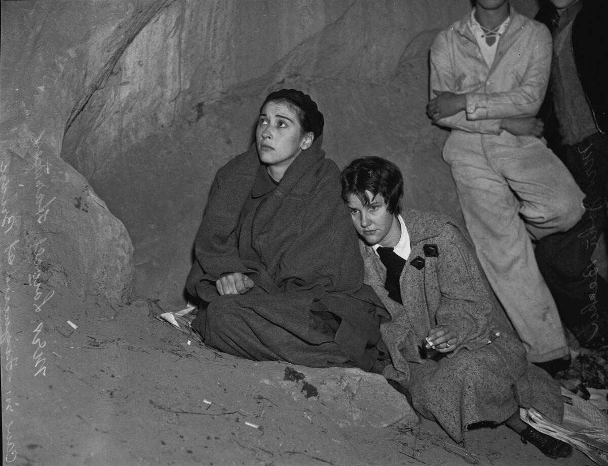 Two young women, one wrapped in a blanket and one smoking a cigarette, huddle together on rocky ground.  