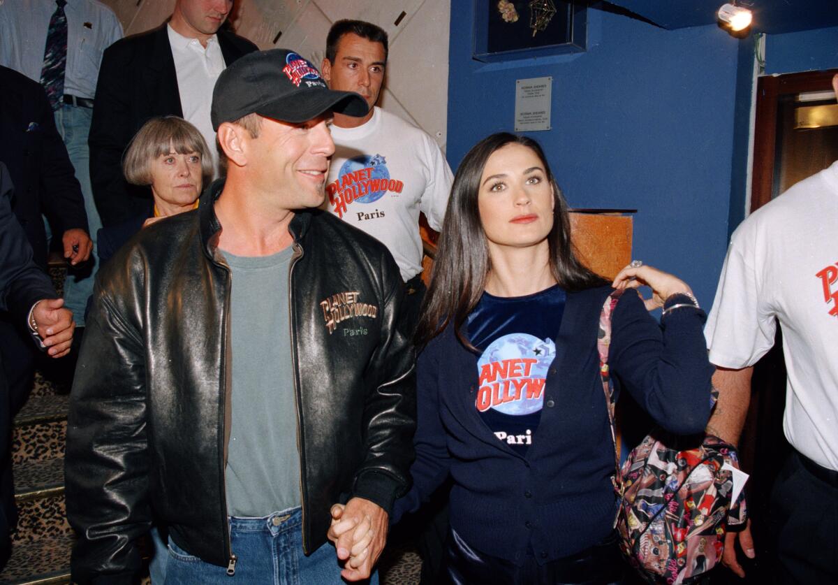 An old image of Bruce Willis in a hat and leather jacket holding hands with Demi Moore in a black shirt