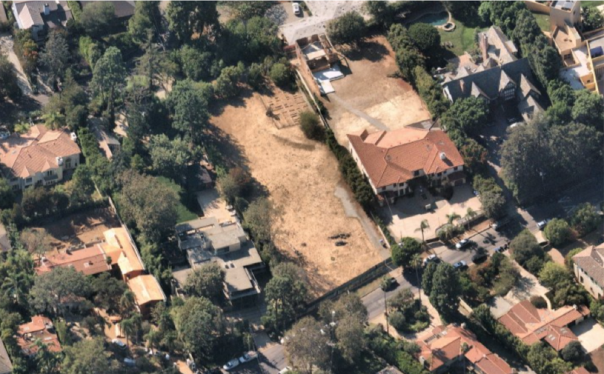 An aerial view of a dirt lot amid developed parcels with large homes and trees.
