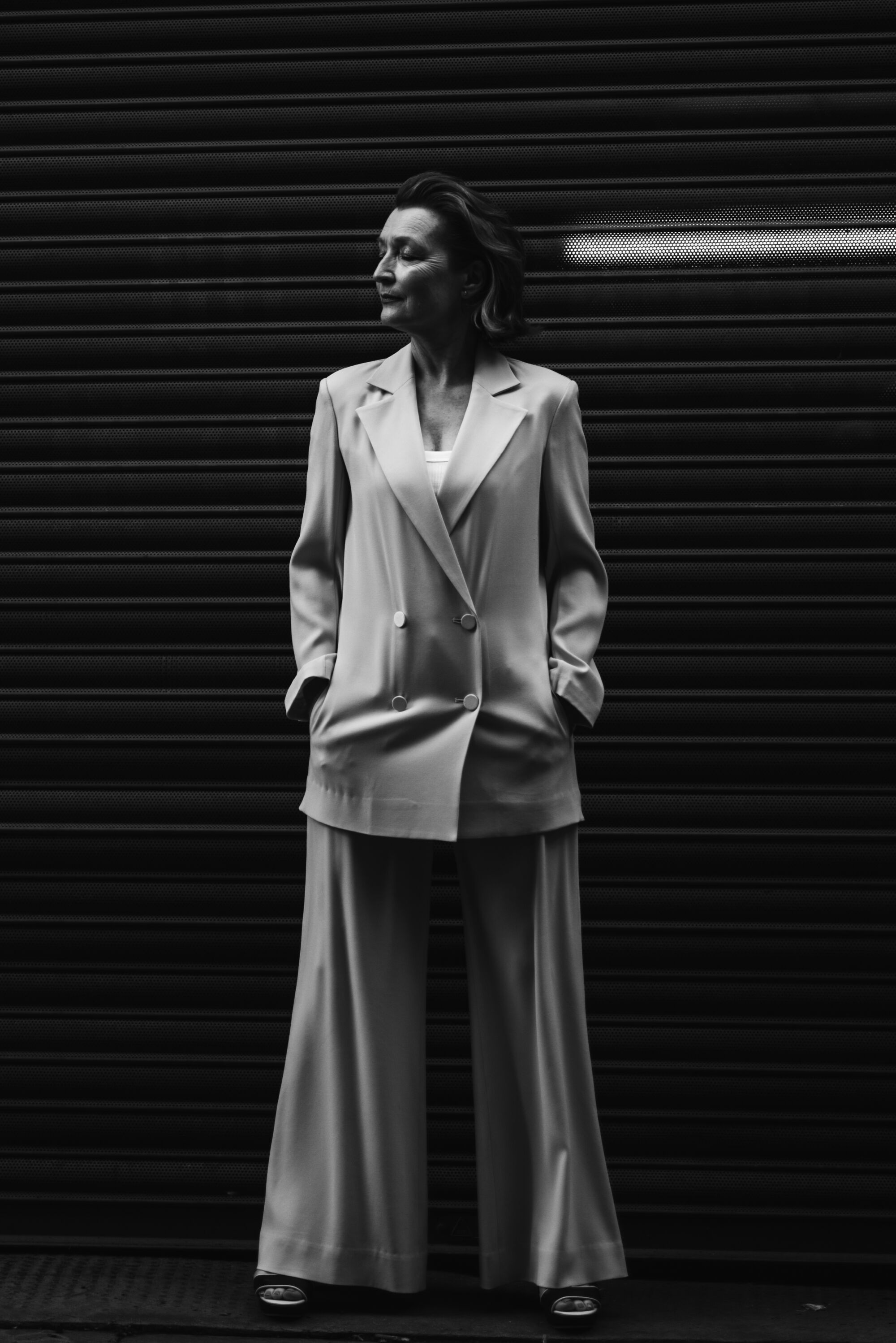 A woman in a white suit leans against a metal garage door closing her eyes with her hands in her jacket pockets 