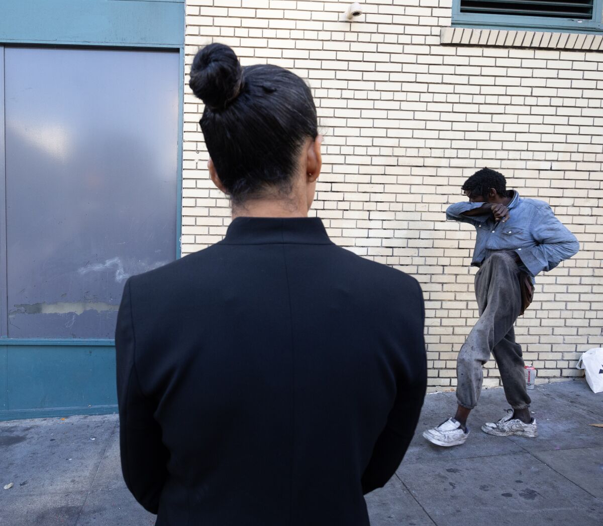 A woman with her hair in a bun observes a homeless person walking on a sidewalk
