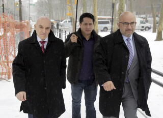 A man in dark clothes is flanked by two others in suits, ties and overcoats in a snow-covered area