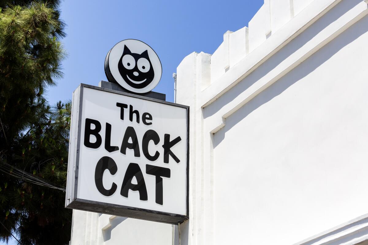 A black and white sign hanging on the side of a white building reads: The Black Cat