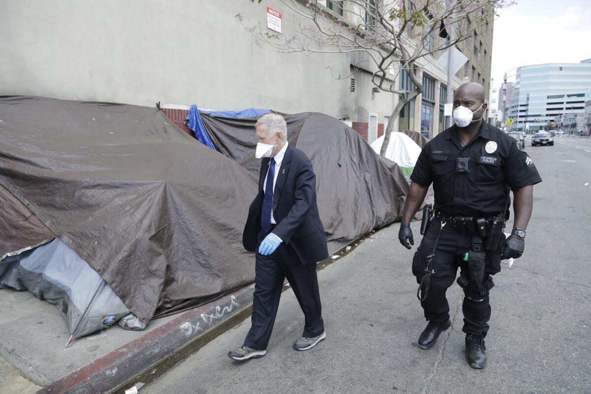 Judge David O. Carter, in a suit and walking shoes, walks next to a police escort past tents and tarps on a sidewalk