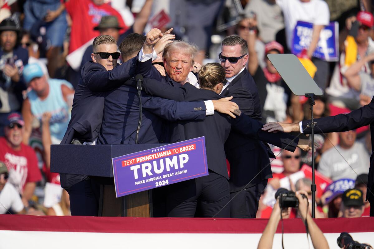 Former President Trump raises a fist as Secret Service agents surround him onstage, with spectators in the stands behind them