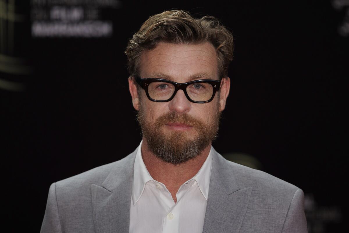 Simon Baker in white button down shirt, gray suit jacket and thick horn-rimmed glasses.