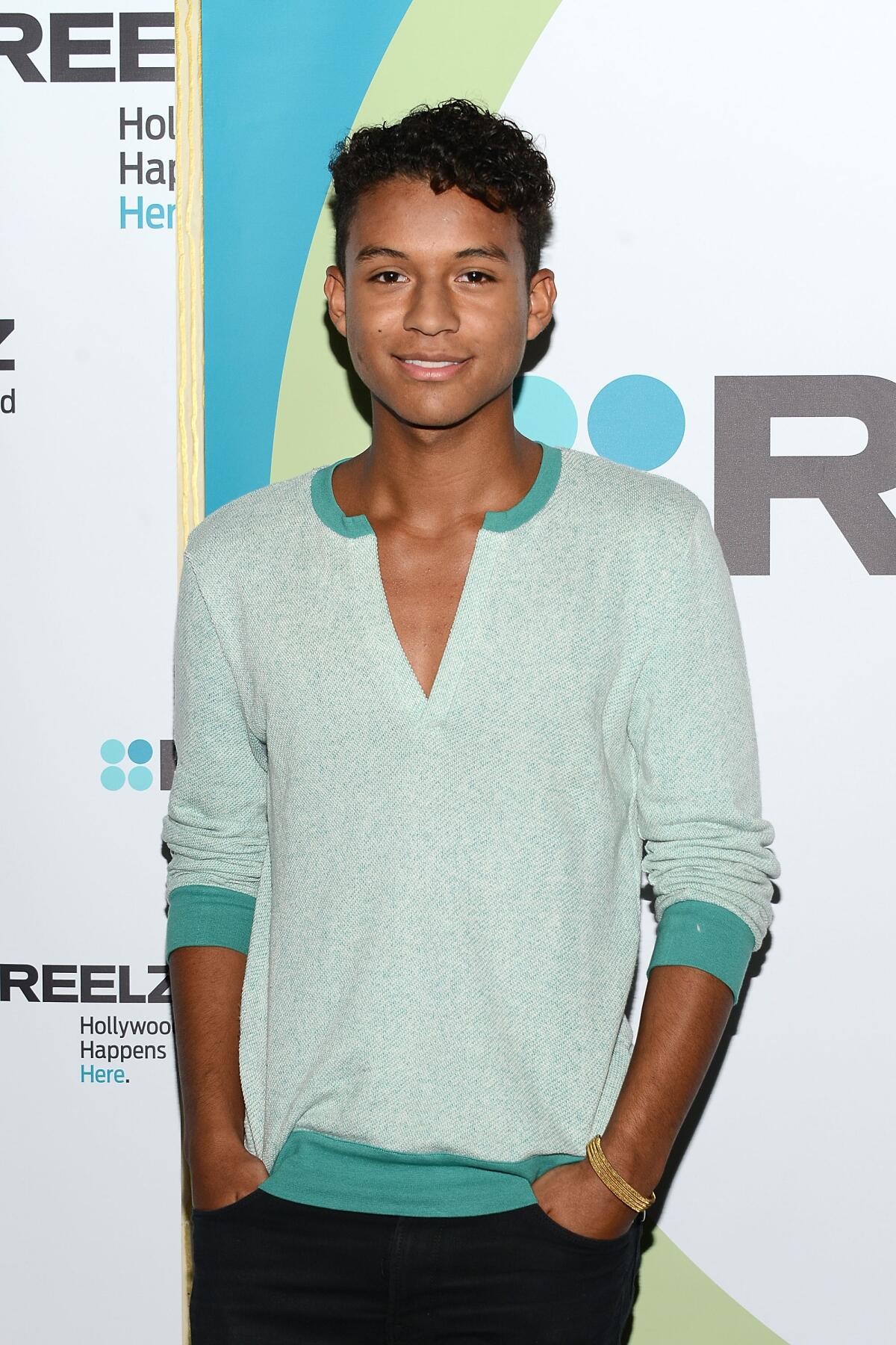 A young man in black pants and a light cool-green v-neck shirt posing for the camera, smiling.