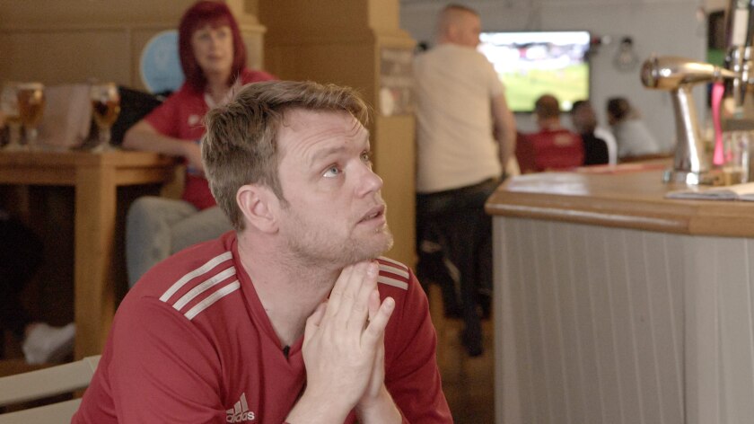A man in a soccer uniform pleads for his team while looking up at a TV screen in a pub.