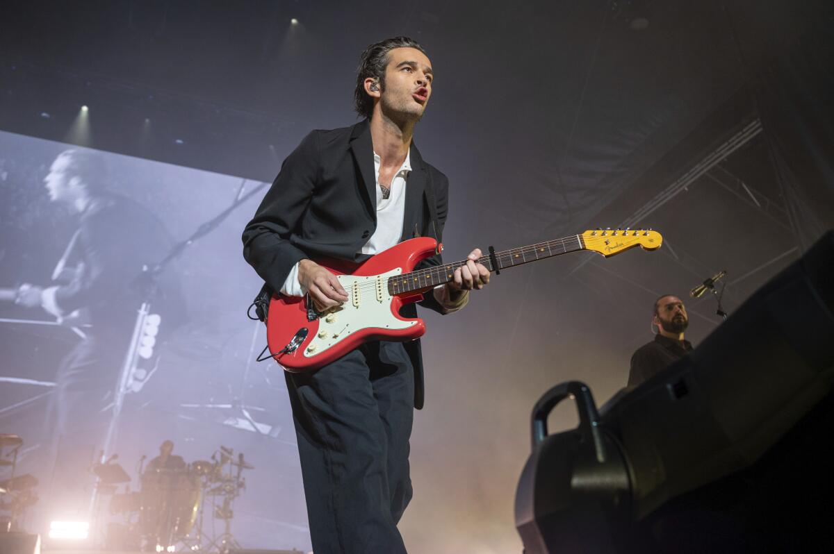 Matty Healy wears a black suit and white shirt as he plays a red-and-white guitar onstage