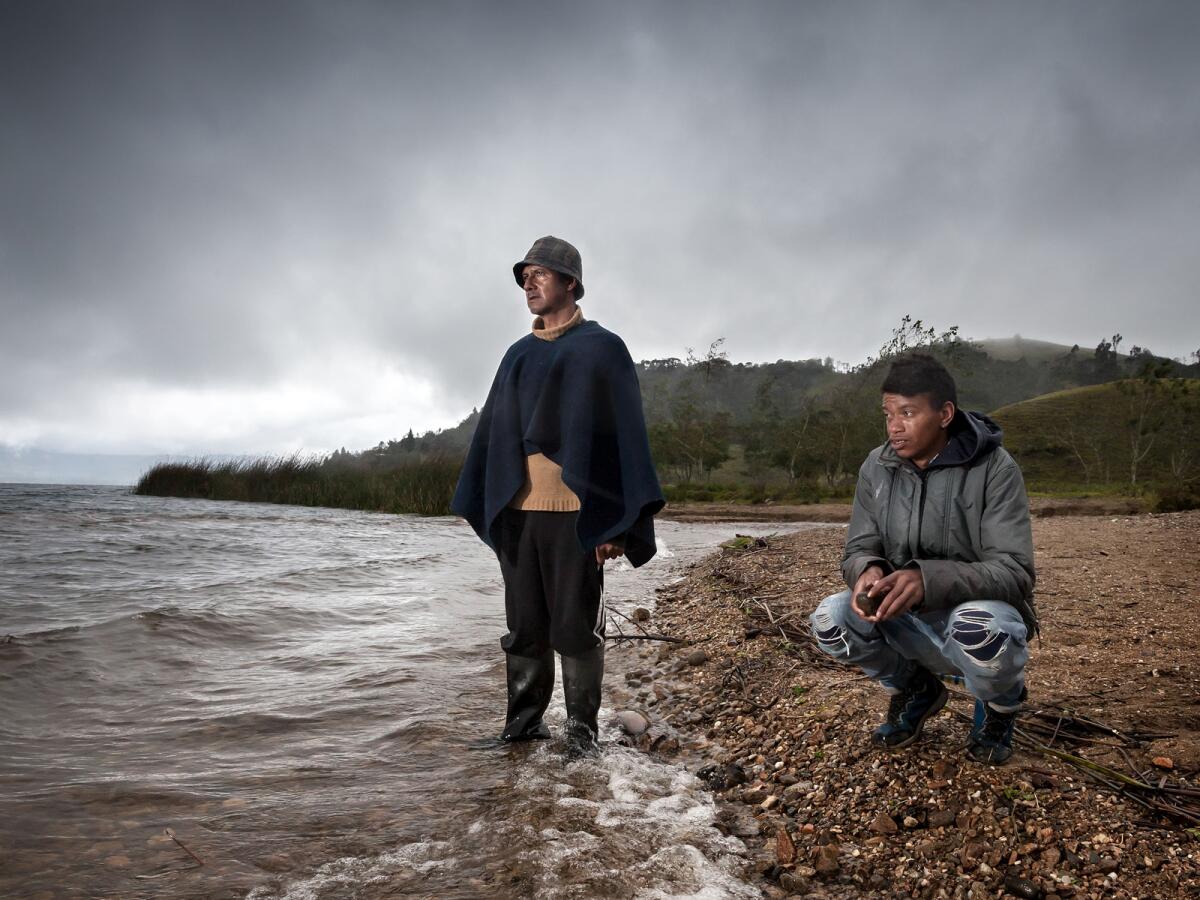 A young man in ripped jeans crouches on a rocky ocean shore alongside a man in wading boots standing in the water.