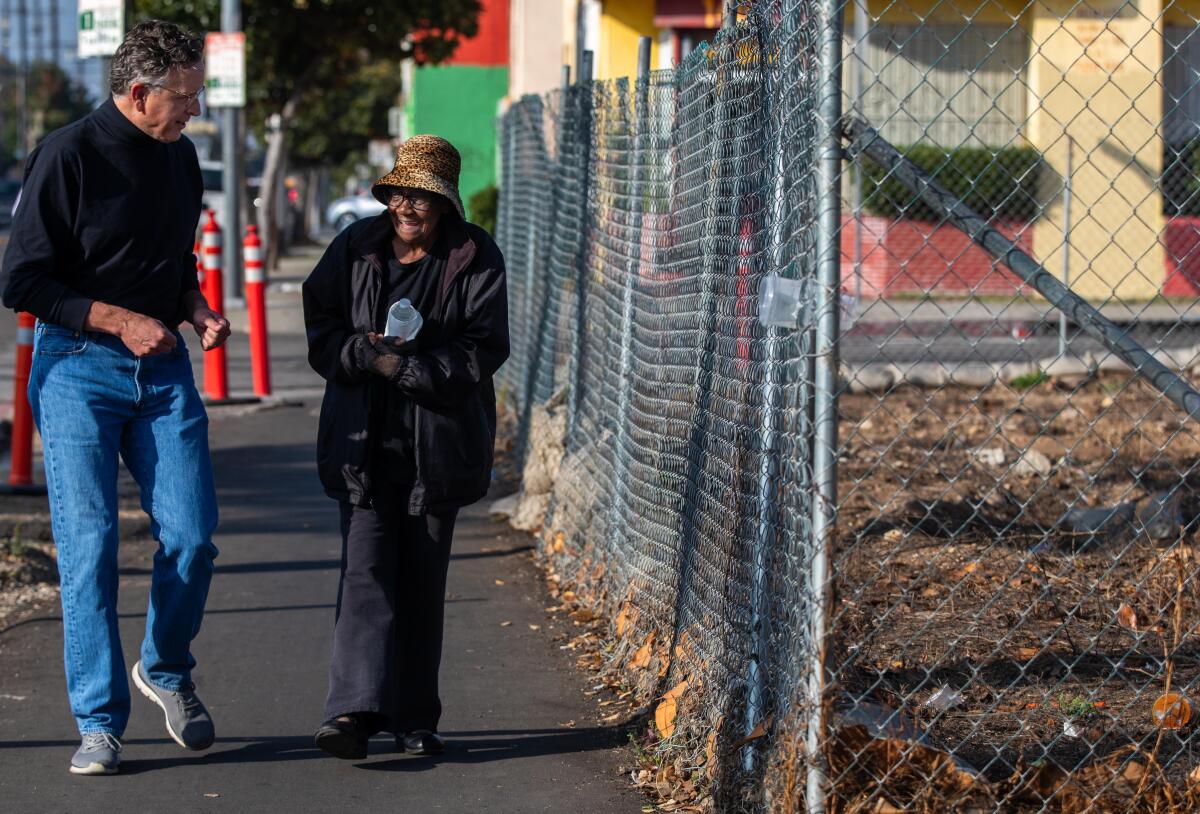A man in jeans and a black shirt, left, and a woman wearing a brown hat and a black jacket walk next to a chainlink fence.