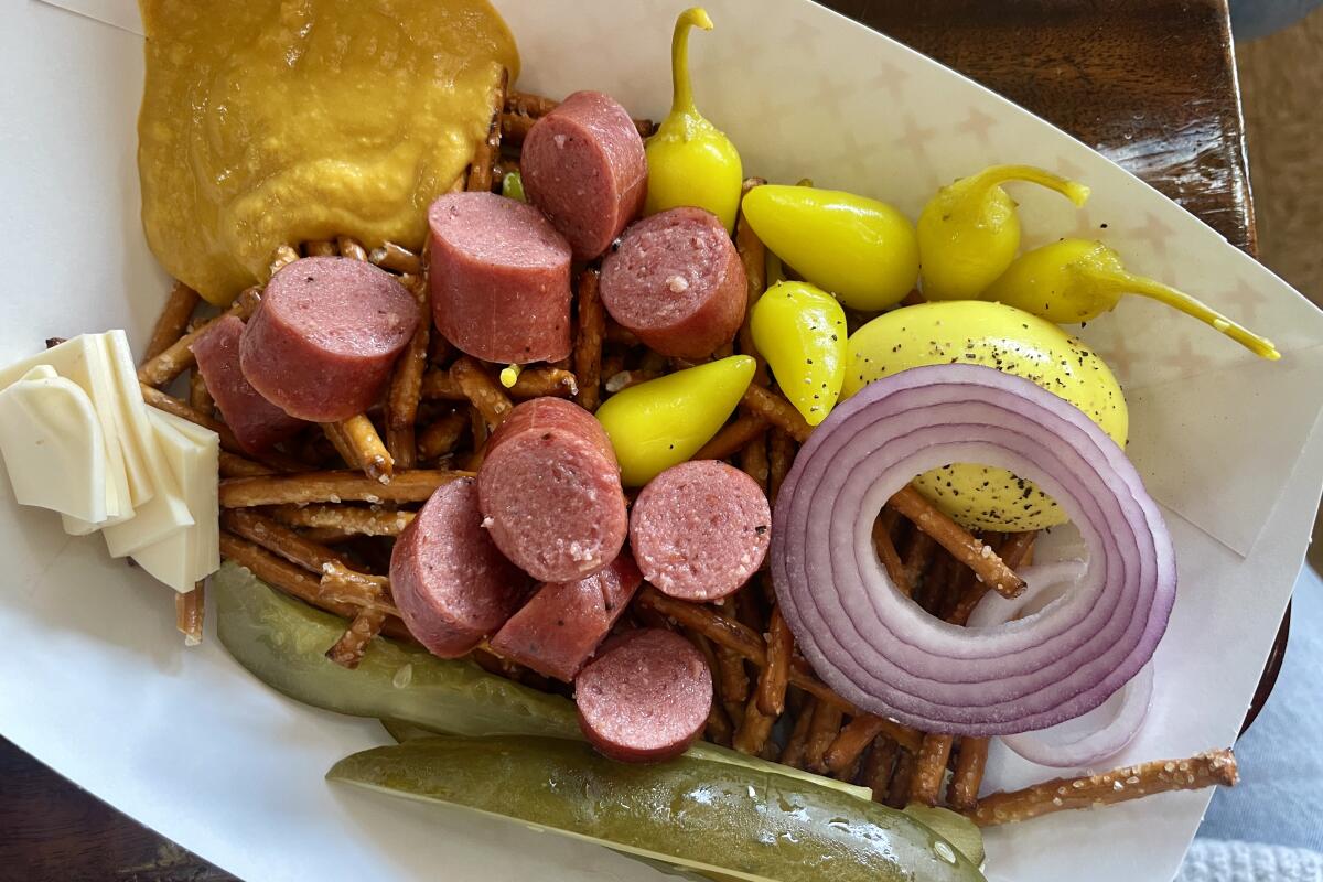 A cardboard boat of sliced sausage, pickle spears, red onion, peppers, mustard, cheese and pretzels