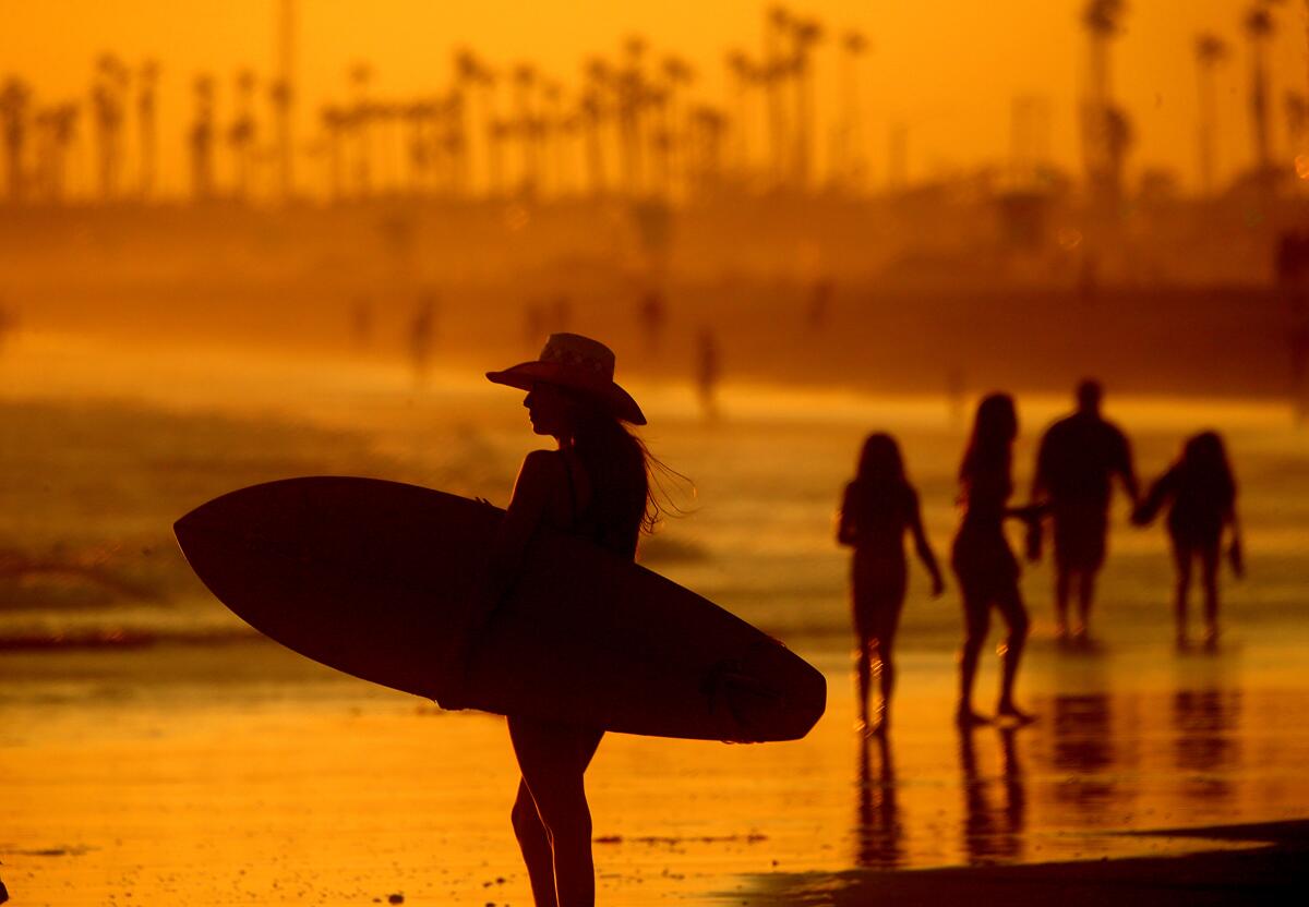 A woman wearing a hat and carrying a surfboard is silhouetted at the beach.