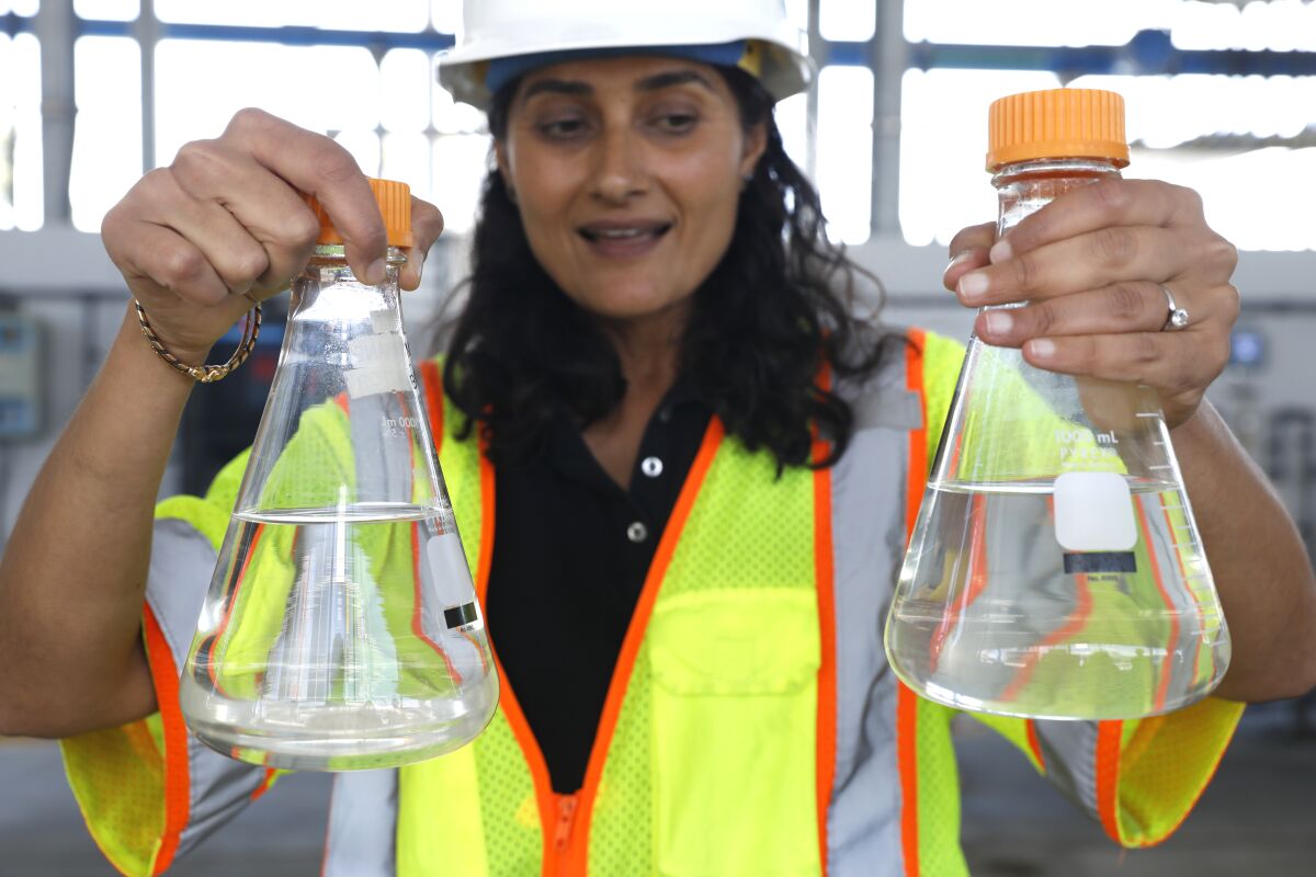 A worker in a hardhat and safety vest displays two conical flasks with clear liquid.