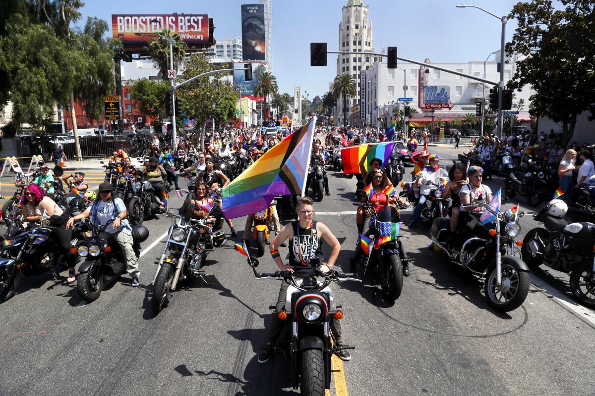 An elevated frame of a street stretching down to buildings on its horizon, with people on their motorcycles with Pride flags
