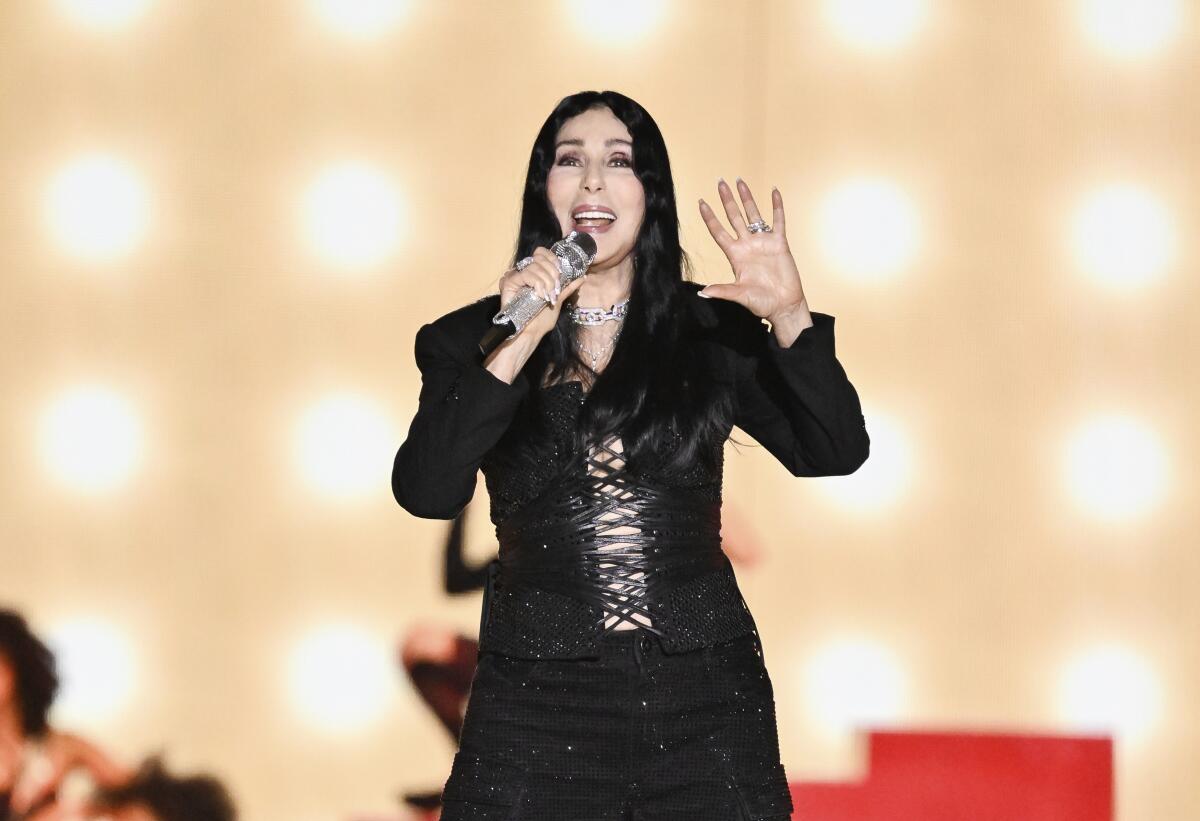 Cher sings into a microphone while holding up one hand in front of a lighted background