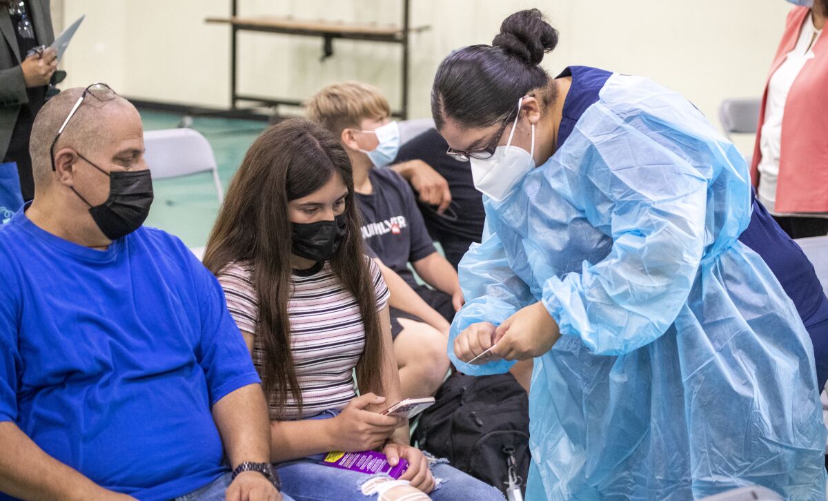 A woman in protective gear speaks with a girl and a man, seated in folding chairs and wearing masks.