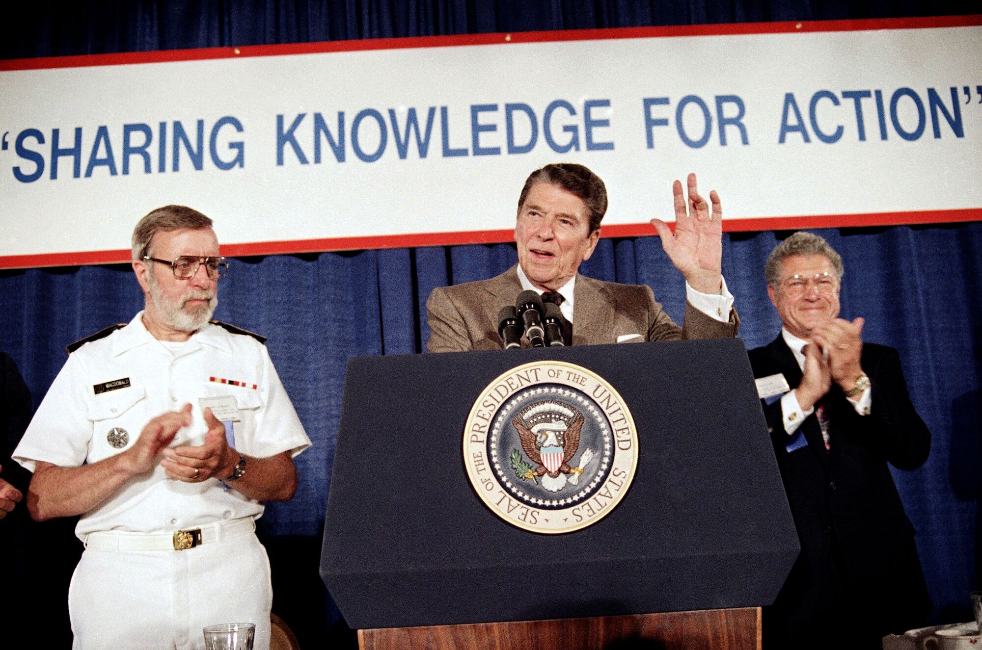 President Reagan speaks at a podium, flanked by two other men