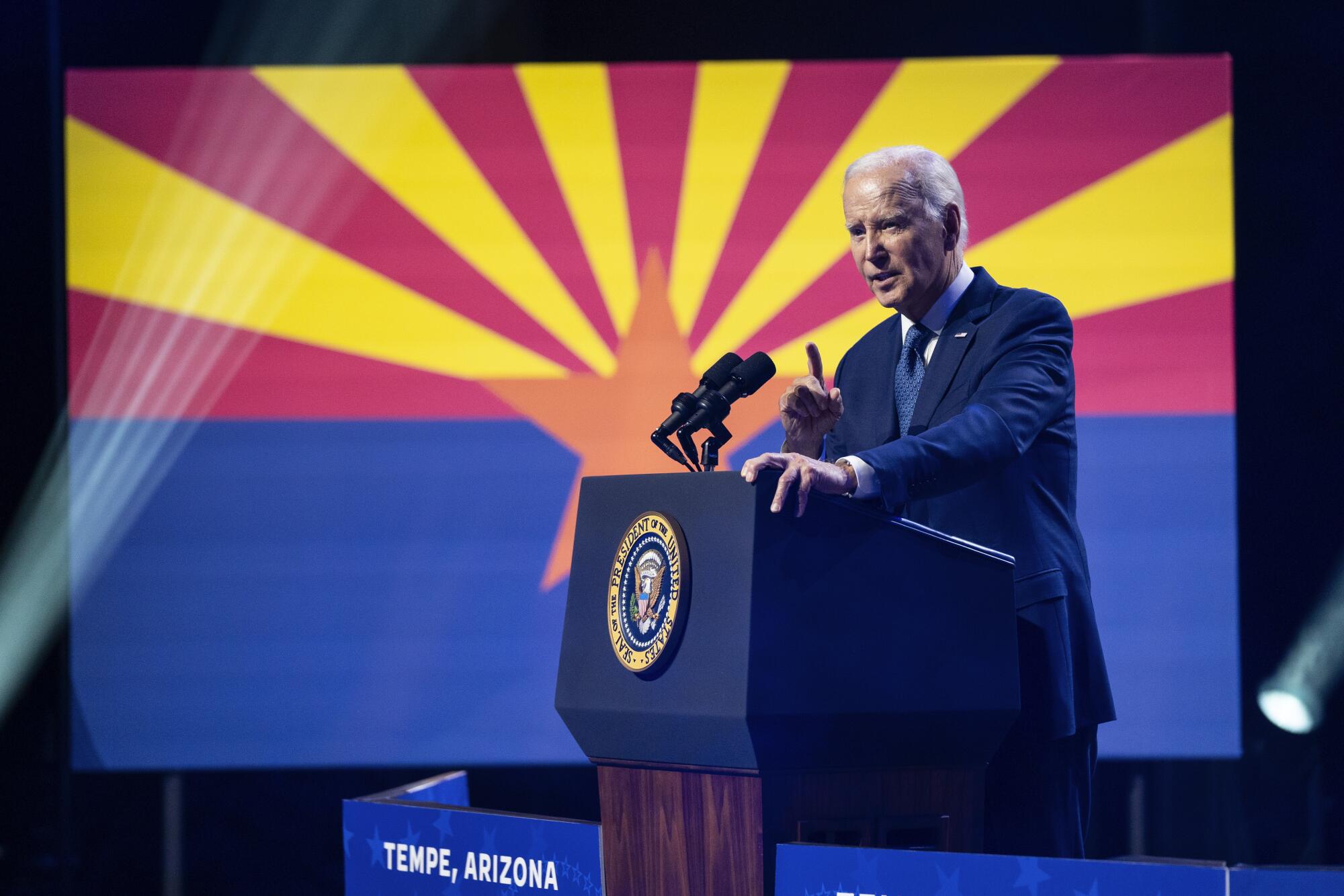 President Biden speaking at a lectern with a presidential seal as Arizona's flag is displayed on a large screen