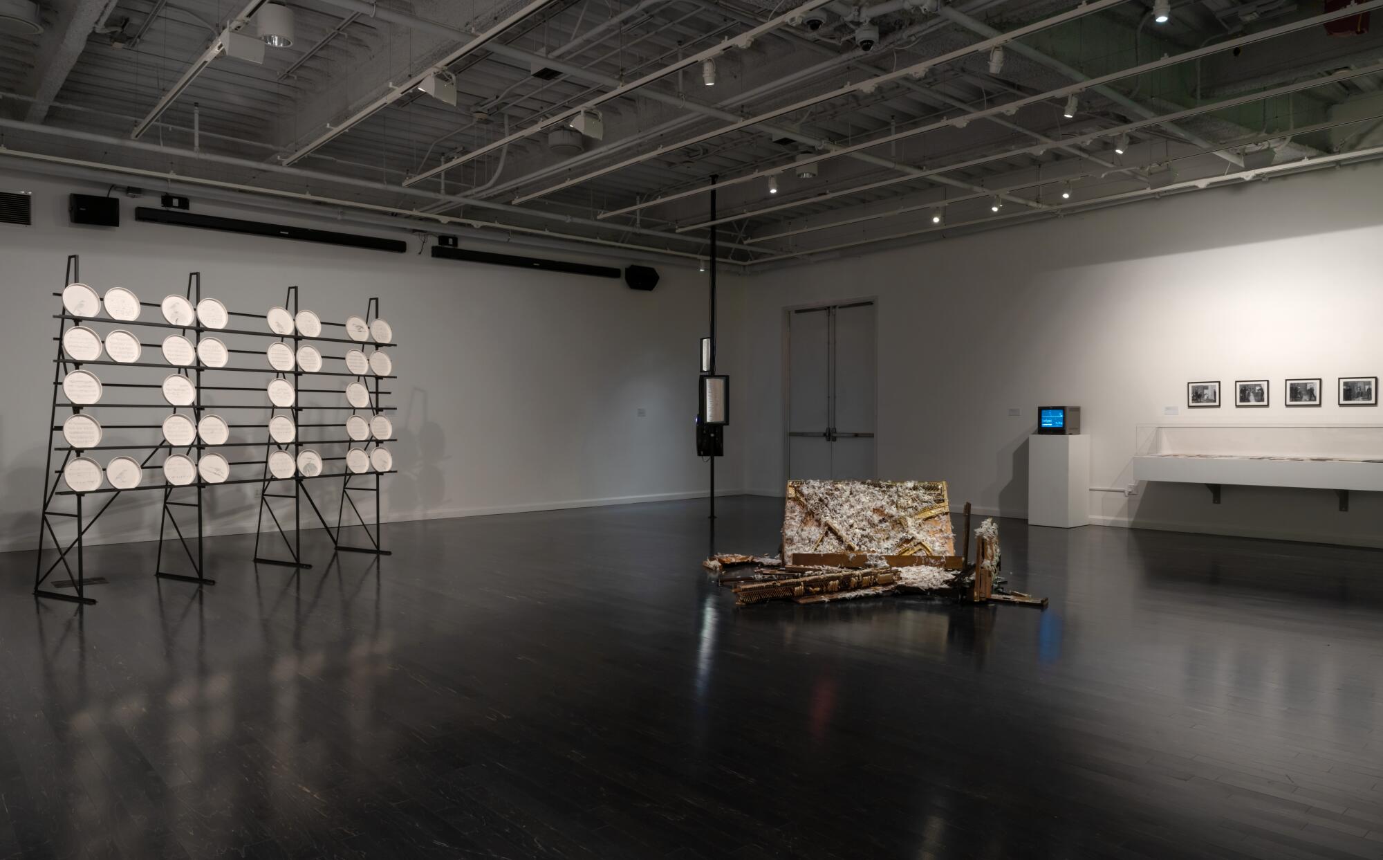 A gallery view shows ceramic disks on a rack, a broken piano and ephemera in a vitrine next to a TV monitor.