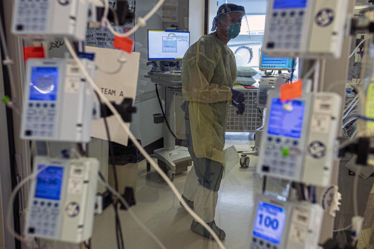 A nurse in full protective gear is seen through a glass door, framed by electronic medical instruments on poles