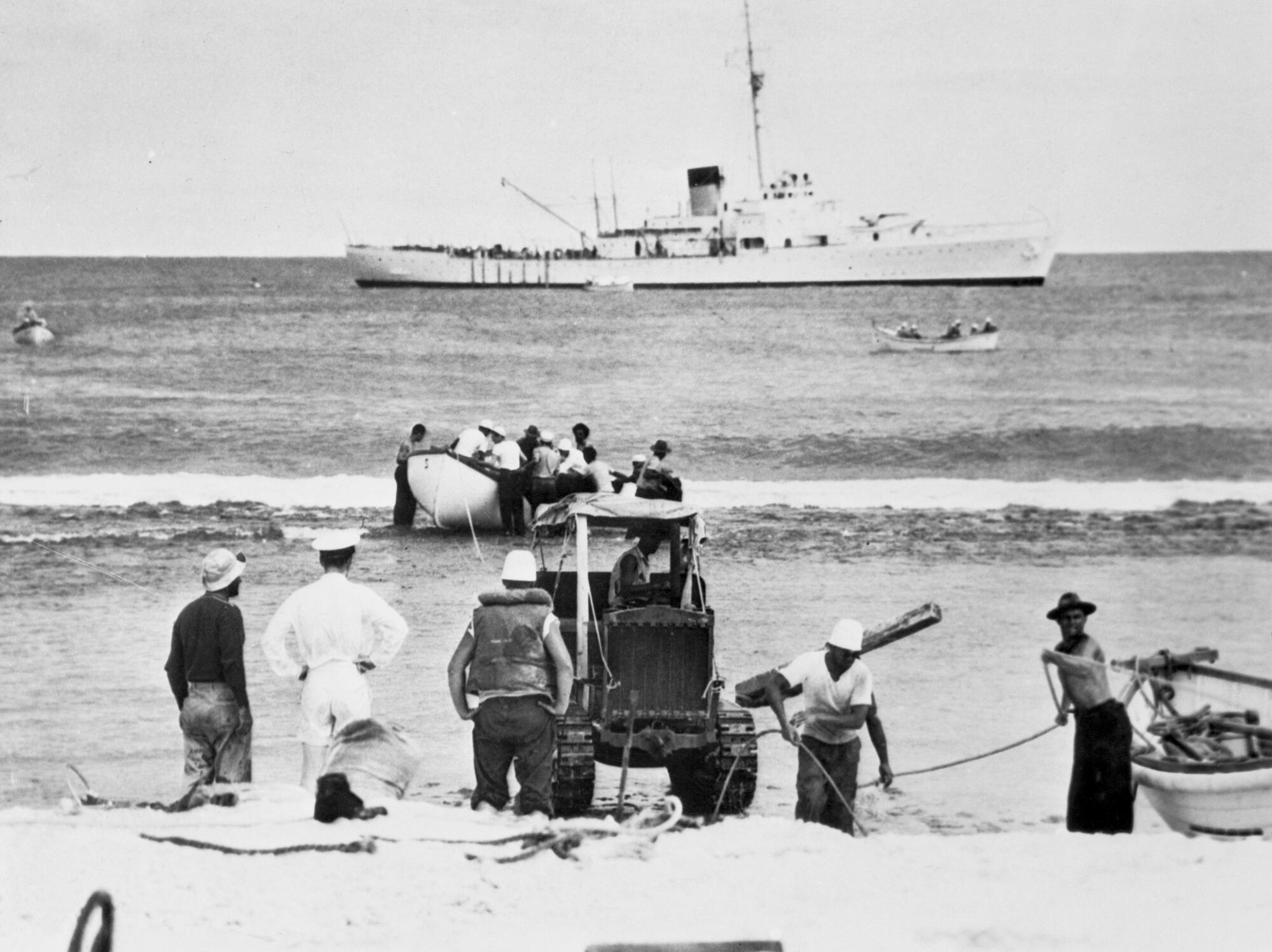 People on a beach with a tractor carry equipment as others beach a small boat while a larger ship lies in the background.