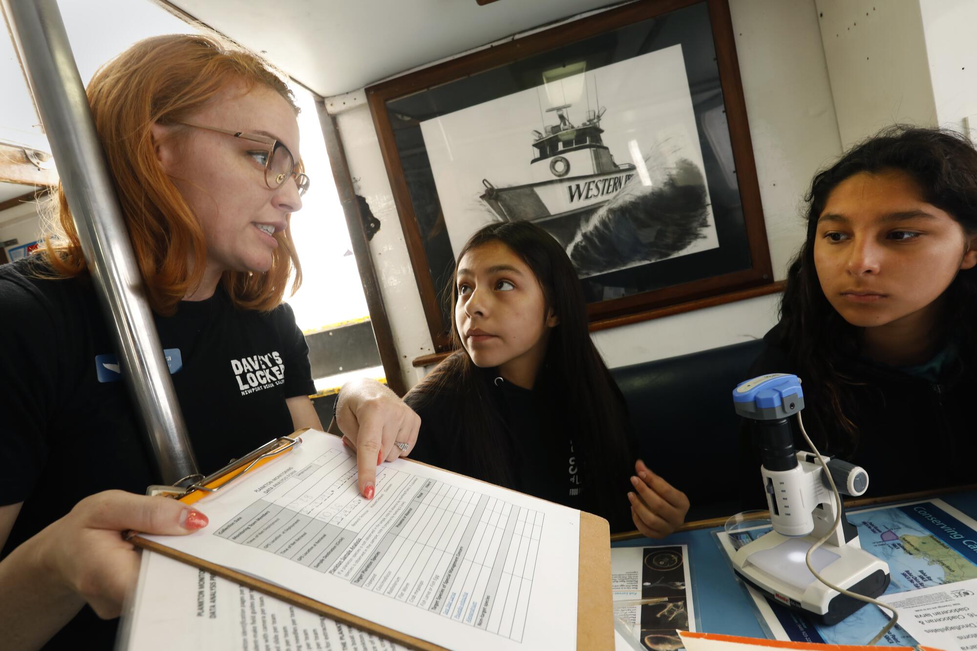 A woman points to a chart on a clipboard and talks with two teenagers next to a small device on a table