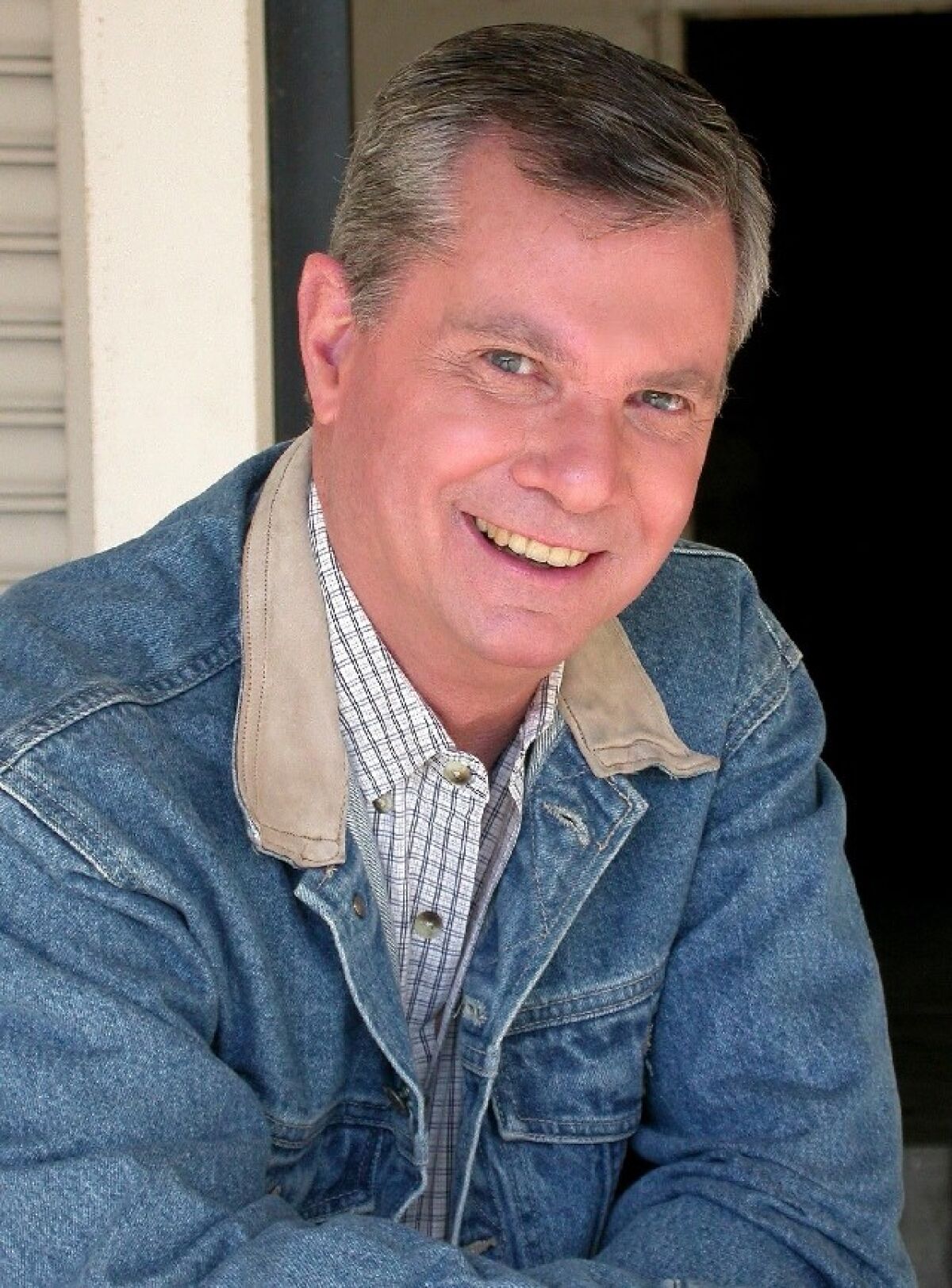 A smiling man in a jean jacket and checkered shirt.
