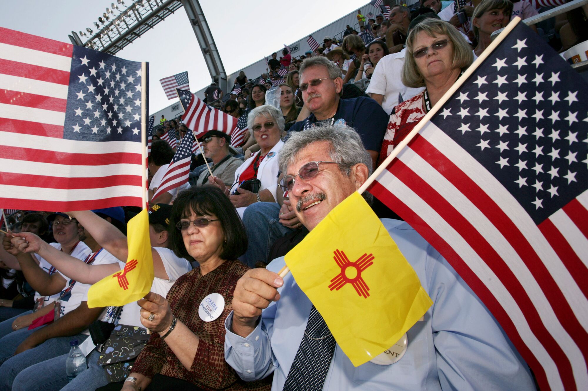 Мужчина, сидящий на трибунах среди толпы, держит звездно-полосатый флаг и красно-желтый флаг Нью-Мексико. 