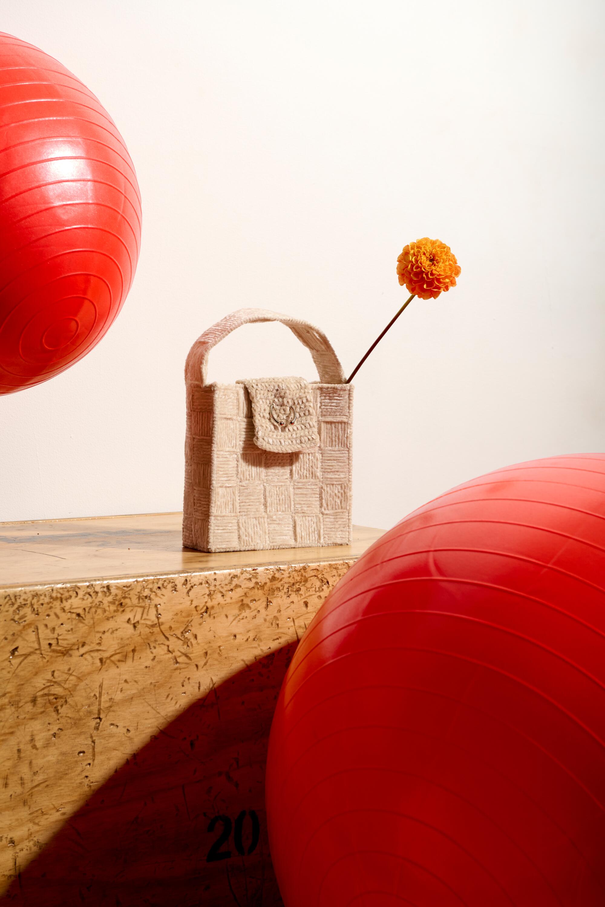 A boxy handbag with a tall-stemmed flower in it sitting on a table near two big red balls.